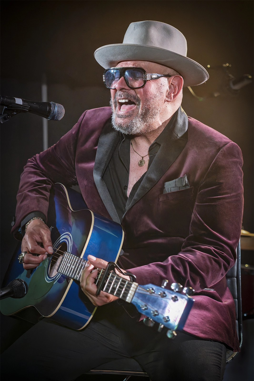 Un musicien aux lunettes noires et chapeau blanc, vêtu d'un blazer bordeaux, chante devant un microphone en jouant d'une guitare acoustique bleue sur scène.
Didier Vaneesbeck