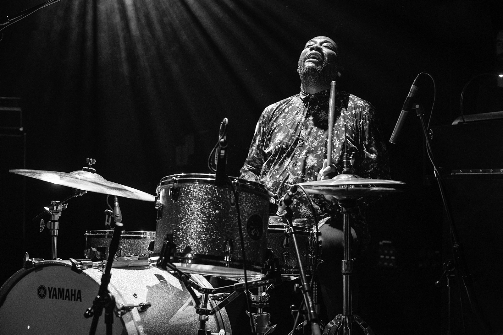 Un batteur  joue de la batterie Yamaha sur scène dans l'obscurité, tête renversée en arrière, concentré dans son jeu musical.
Didier  Vaneesbeck