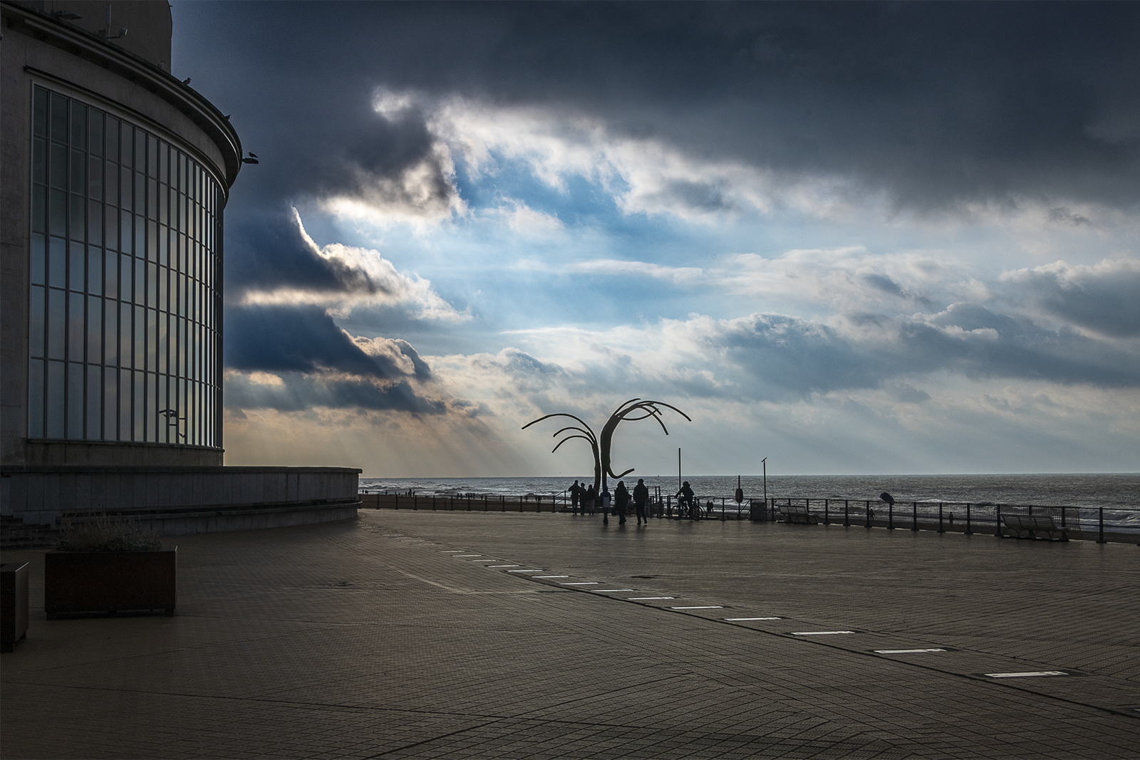 Photographie prise à la côte belge montrant une installation métallique sculpturale aux formes courbes qui s'élèvent vers un ciel dramatique nuageux, avec un bâtiment moderne à gauche et quelques visiteurs en contrebas, capturée par le photographe bruxellois Didier Vaneesbeck.