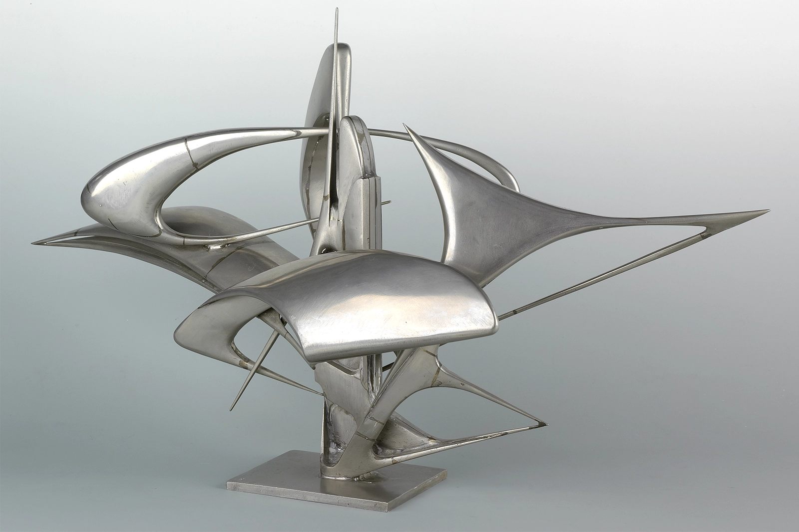 Sculpture en métal argenté aux formes géométriques épurées et courbes fluides, composée de plusieurs éléments pointus et arrondis assemblés verticalement sur une base plate, photographiée sur fond gris neutre.