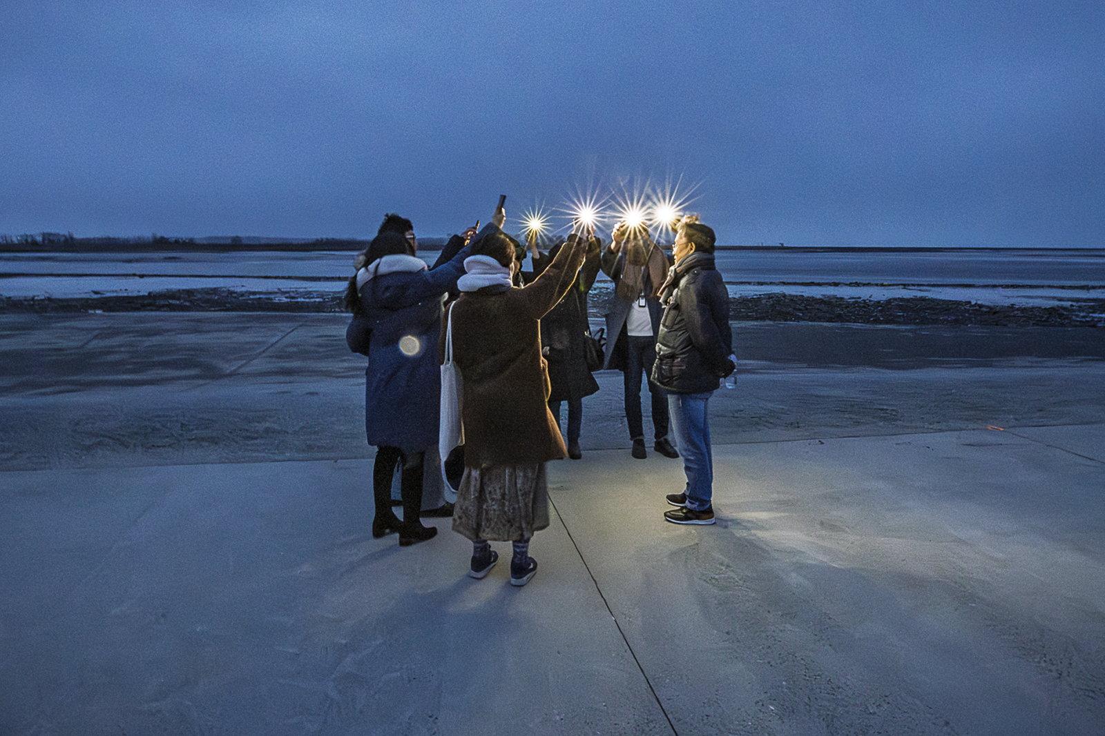 Un groupe de personnes se tient en cercle sur une plage gelée au crépuscule, les bras levés tenant des sources lumineuses qui créent des étoiles de lumière au-dessus de leurs têtes, sous un ciel bleu nuit. Didier Vneesbeck