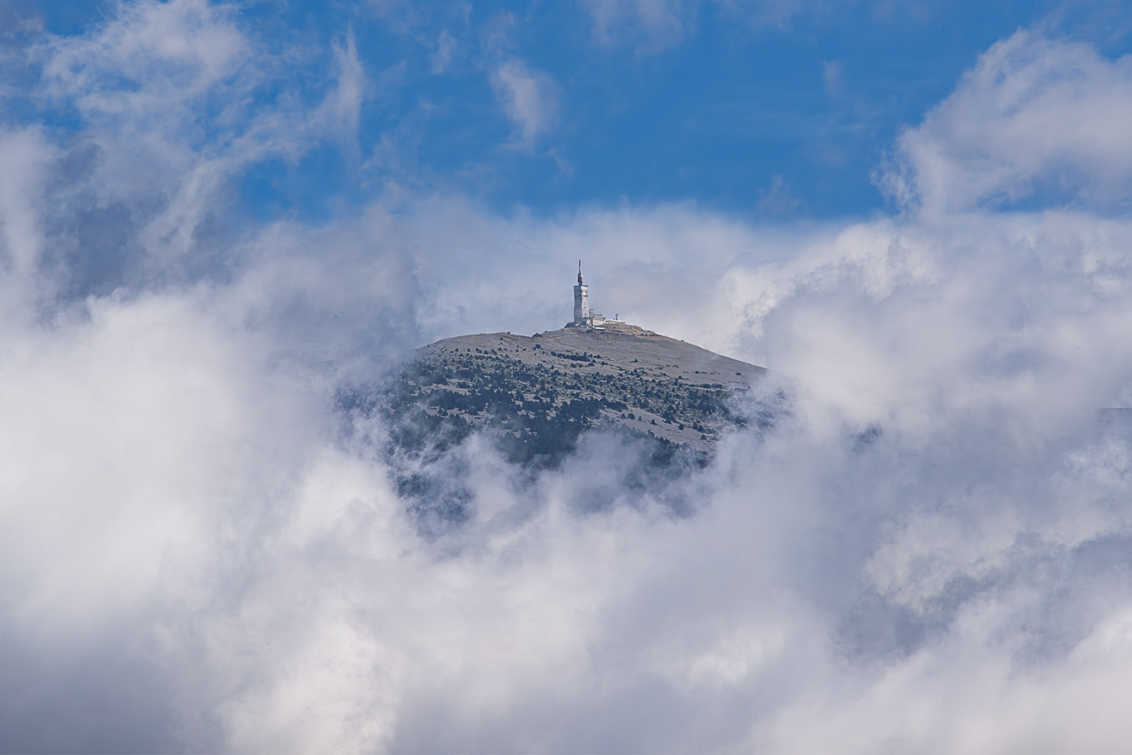 Montagne rocheuse couronnée d'une basilique blanche émergeant partiellement des nuages épais, sous un ciel bleu clair parsemé de formations nuageuses.
Didier Vaneesbeck