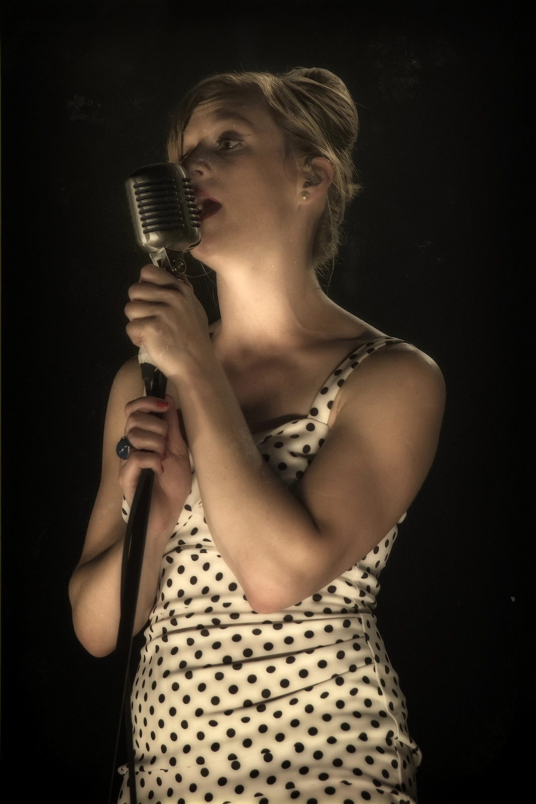 Une femme aux cheveux blonds coiffés en chignon chante passionnément devant un microphone vintage chromé, vêtue d'une robe blanche à pois noirs, sur fond noir. Didier Vaneesbeck