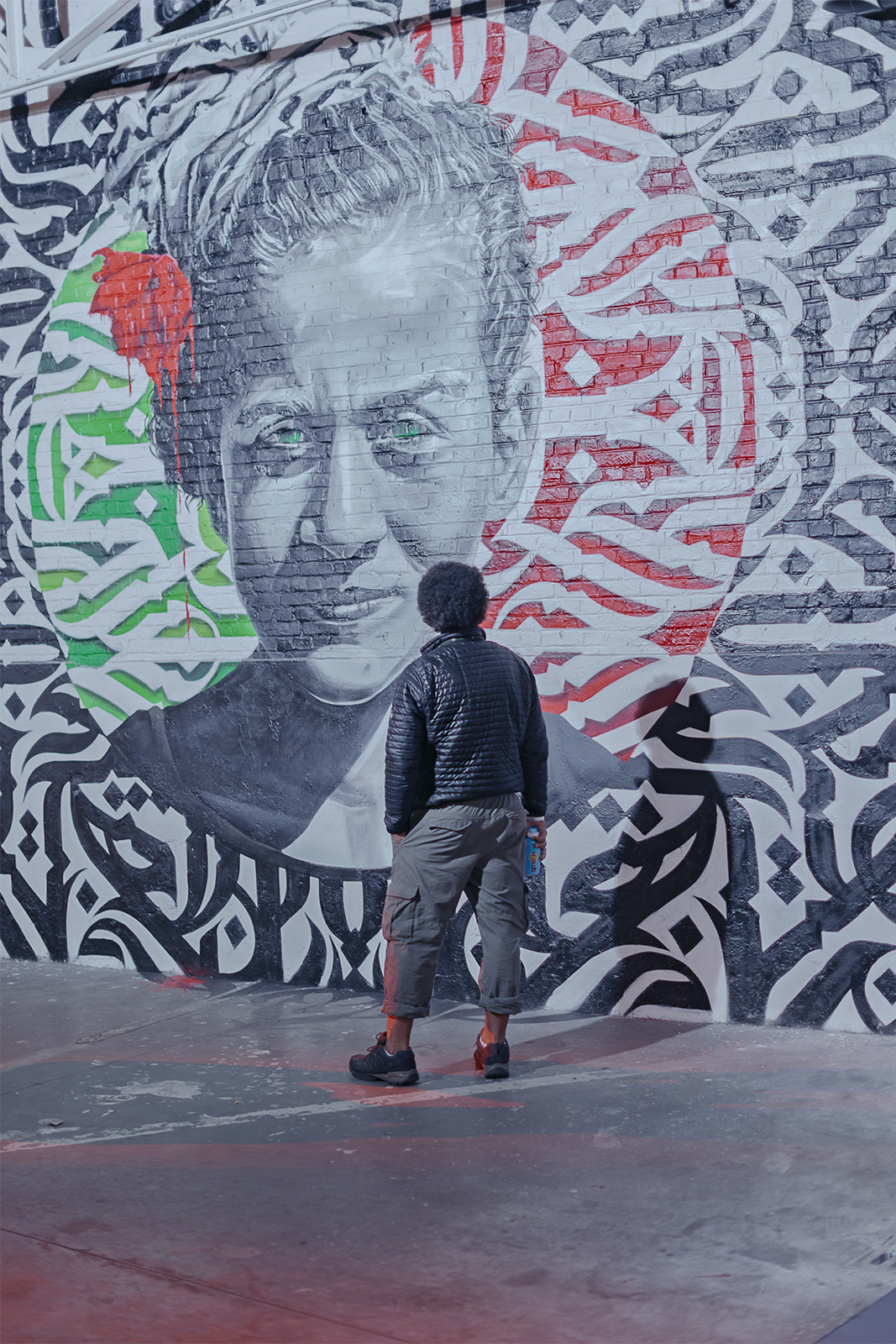 Un visiteur de dos, vêtu d'une veste noire matelassée, observe un imposant portrait mural de street art sur un mur de briques à Bruxelles, composé de motifs géométriques en noir, blanc, rouge et vert avec des yeux verts luminescents.