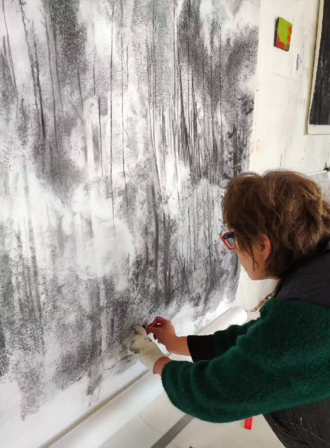 Vue de dos de Véronique Poppe dessinant à la mine de plomb sur une grande feuille blanche tendue au mur, révélant son processus créatif.