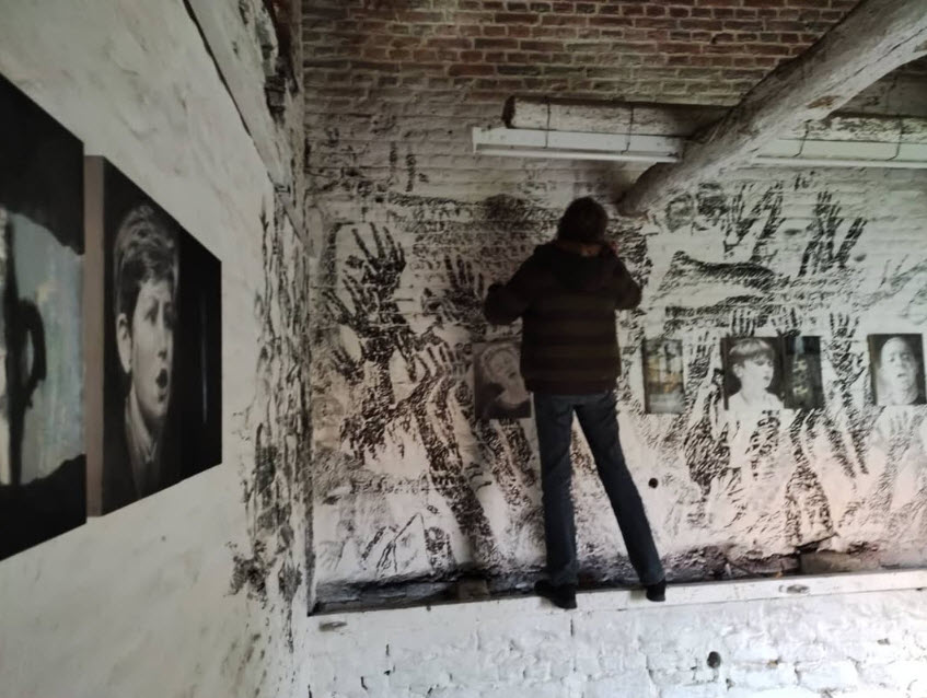 Véronique Poppe vêtue de noir se tient dans un espace industriel aux murs blancs couverts d'empreintes de mains noires et de dessins, avec des portraits photographiques affichés sur la gauche, illustrant une installation ou un projet artistique urbain.