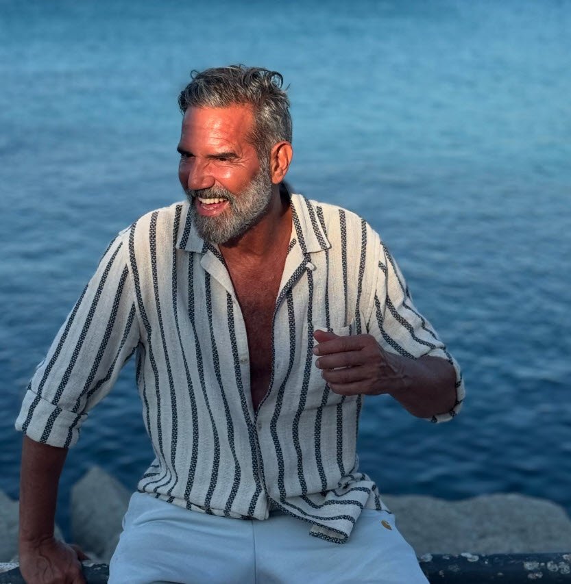 Portrait d'Olivier Figus Lechevalier, comédien, souriant face à la caméra en chemise rayée blanche et bleu marine, assis au bord de l'eau avec la mer en arrière-plan.

Portrait estival d'Olivier Figus Lechevalier - Artiste comédien belge