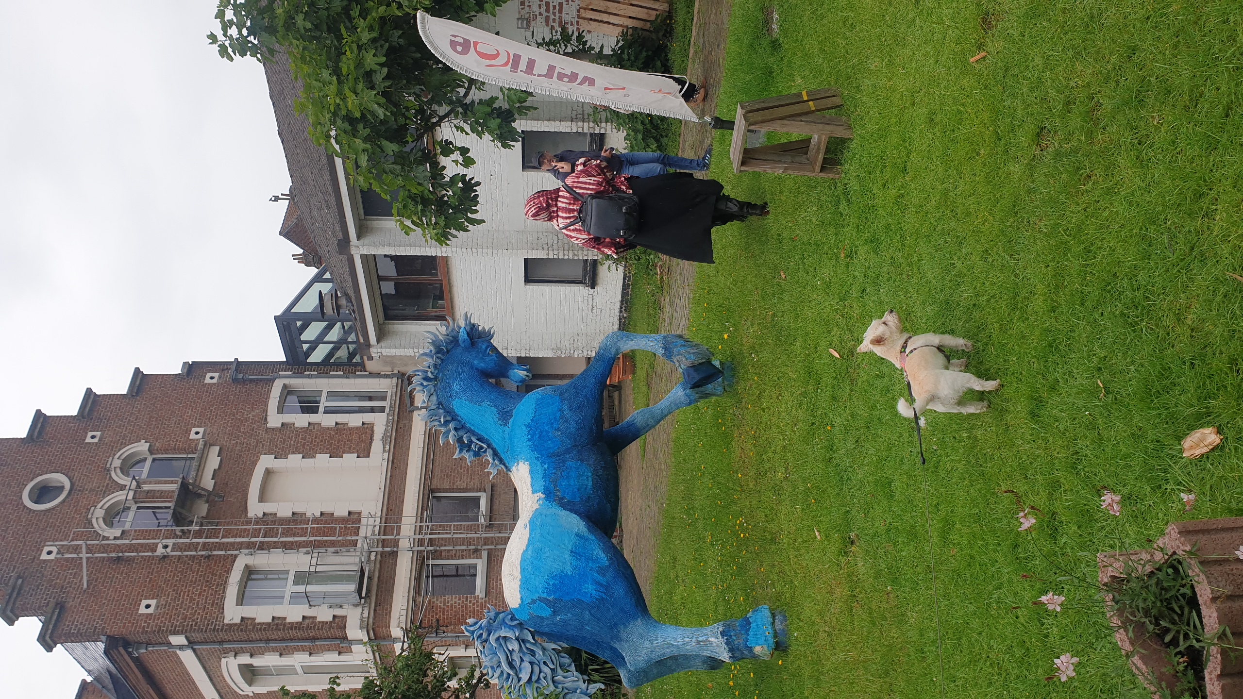 Sculpture d'un cheval bleu vif à crinière hérissée exposée dans un jardin devant un bâtiment historique en briques, avec des visiteurs et un chien en premier plan. Galerie Vertige. Exposition peinture Bruxelles.