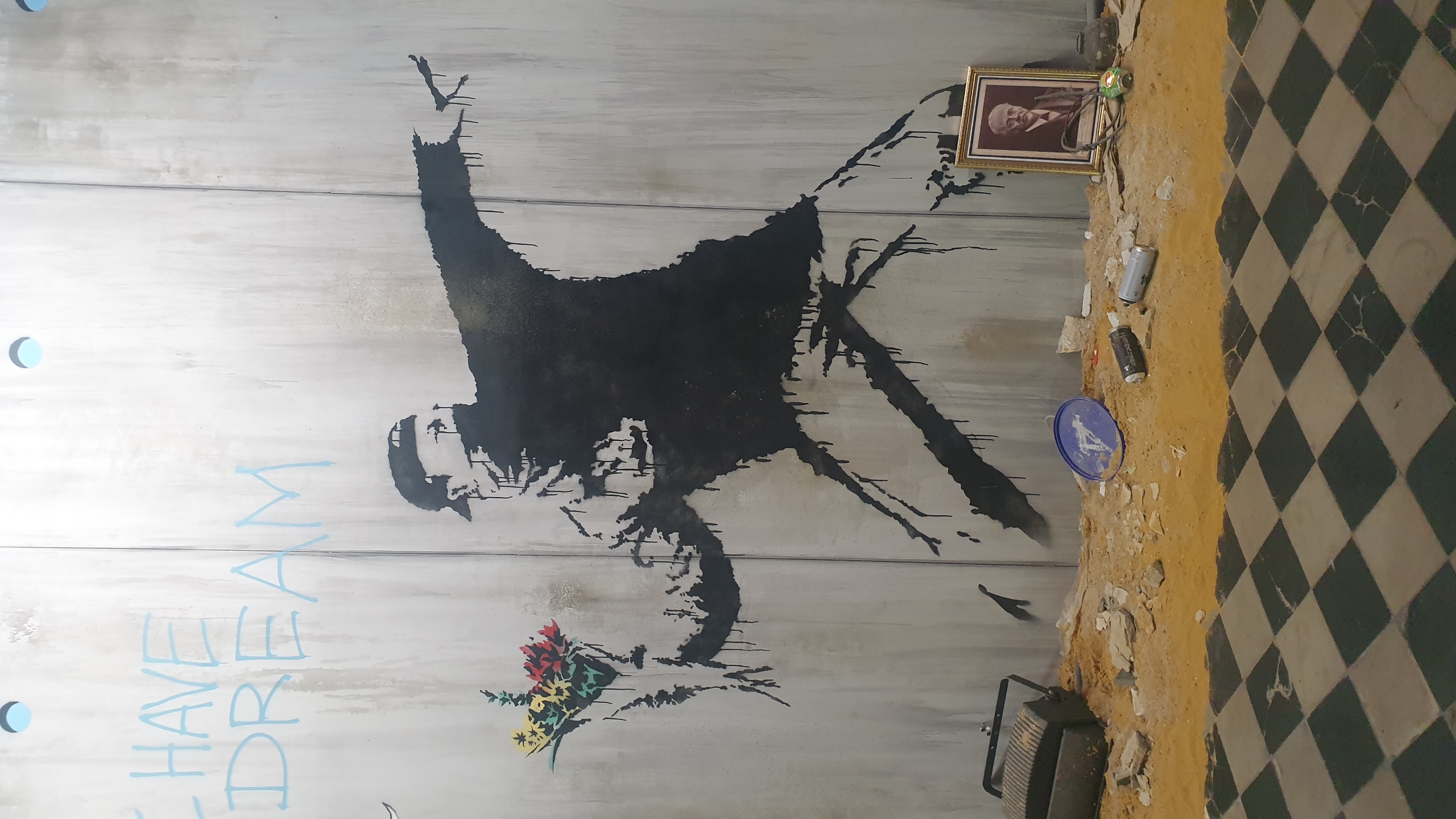 Photographie de street art bruxellois au Musée Banksy Bruxelles sur mur gris : silhouette noire d'une figure dynamique en position de lancer, tenant un bouquet de fleurs multicolores, avec texte bleu « I HAVE A DREAM » au-dessus, entourée d'objets dispersés au sol. By Dropmuse.art https://www.dropmuse.art