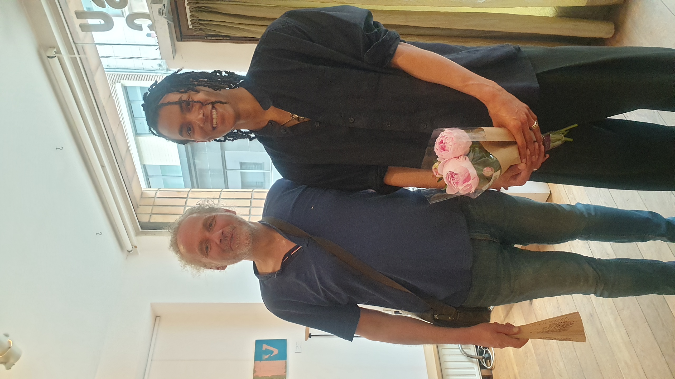 Bailly Tshibangu et Jerome Selosse (Vernissage Artbase 2026) posent ensemble dans un espace de galerie contemporaine à Bruxelles ; celui de droite, artiste du collectif Dropmuse.Art, tient un bouquet de roses roses, tandis que celui de gauche porte un sac en bandoulière.