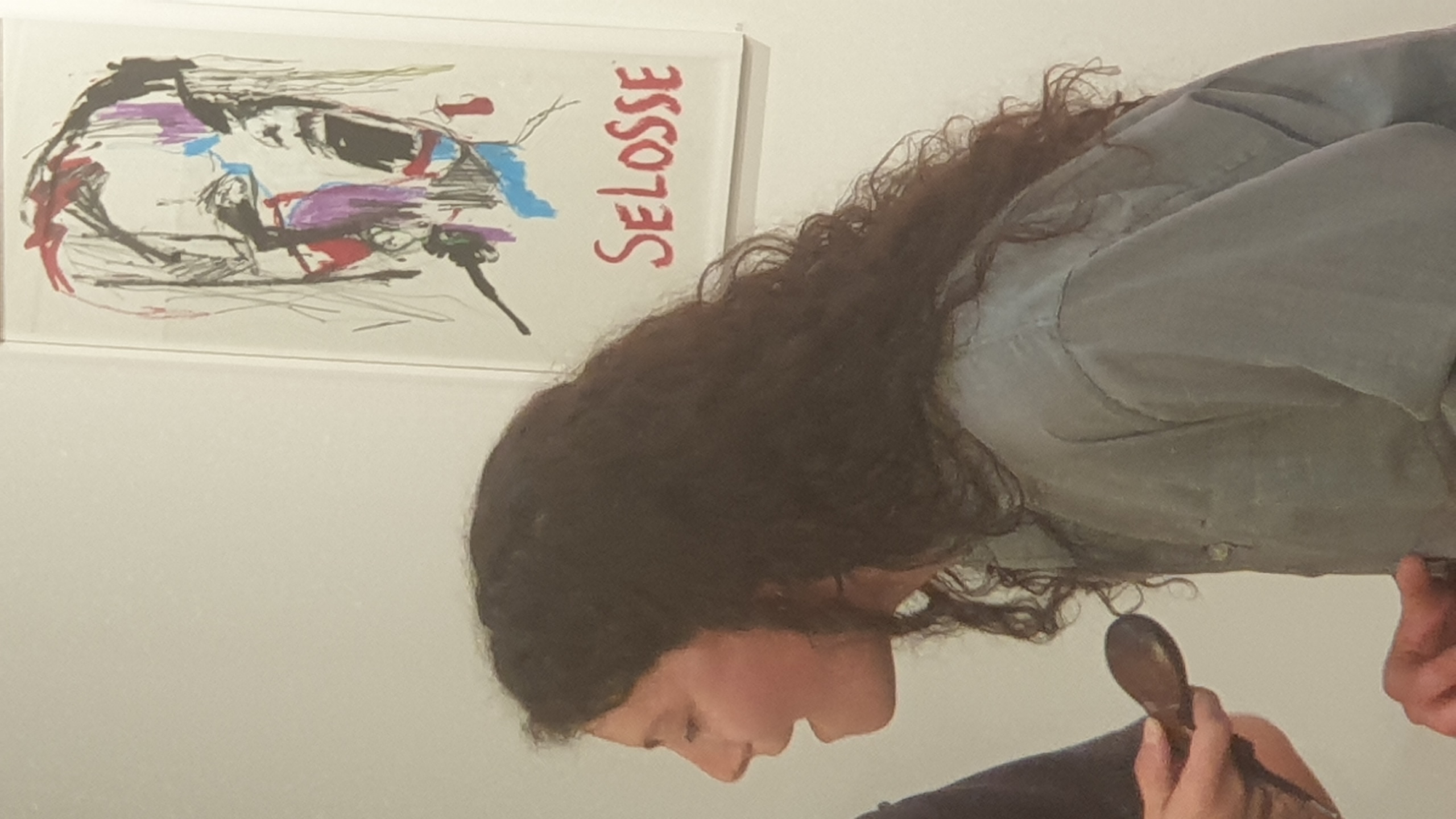 Une femme de profil observe une œuvre d'art accrochée au mur représentant un portrait abstrait en noir avec des touches de couleur rouge, bleu et violet, intitulée 'SELOSSE', dans un espace d'exposition.