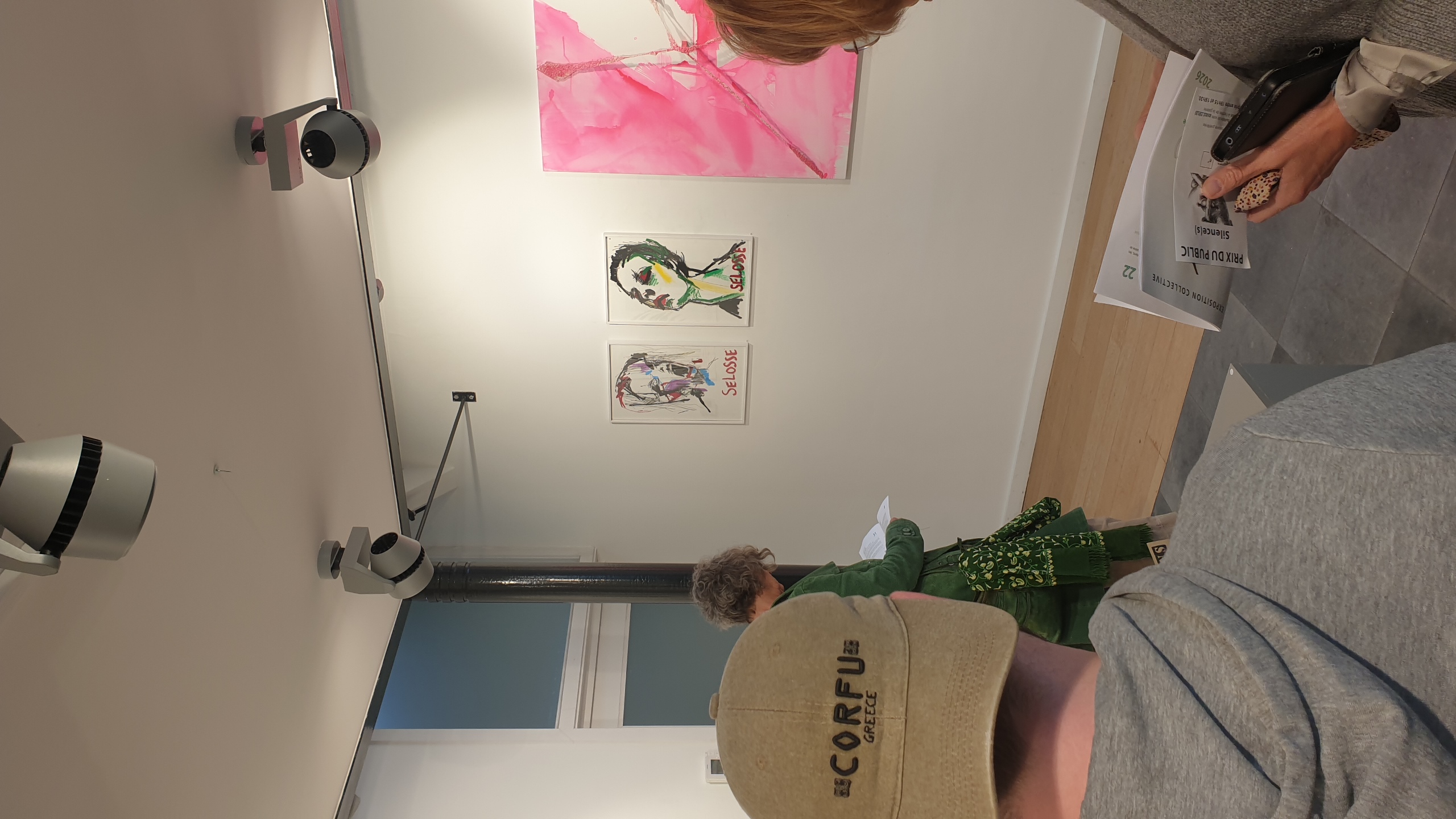Deux visiteurs observent des œuvres d'art accrochées aux murs blancs d'une galerie : des portraits expressionnistes de  « Jérôme SELOSSE » et une grande composition rose fluo, sous éclairage professionnel. Expo Galerie Vertige (Bruxelles)