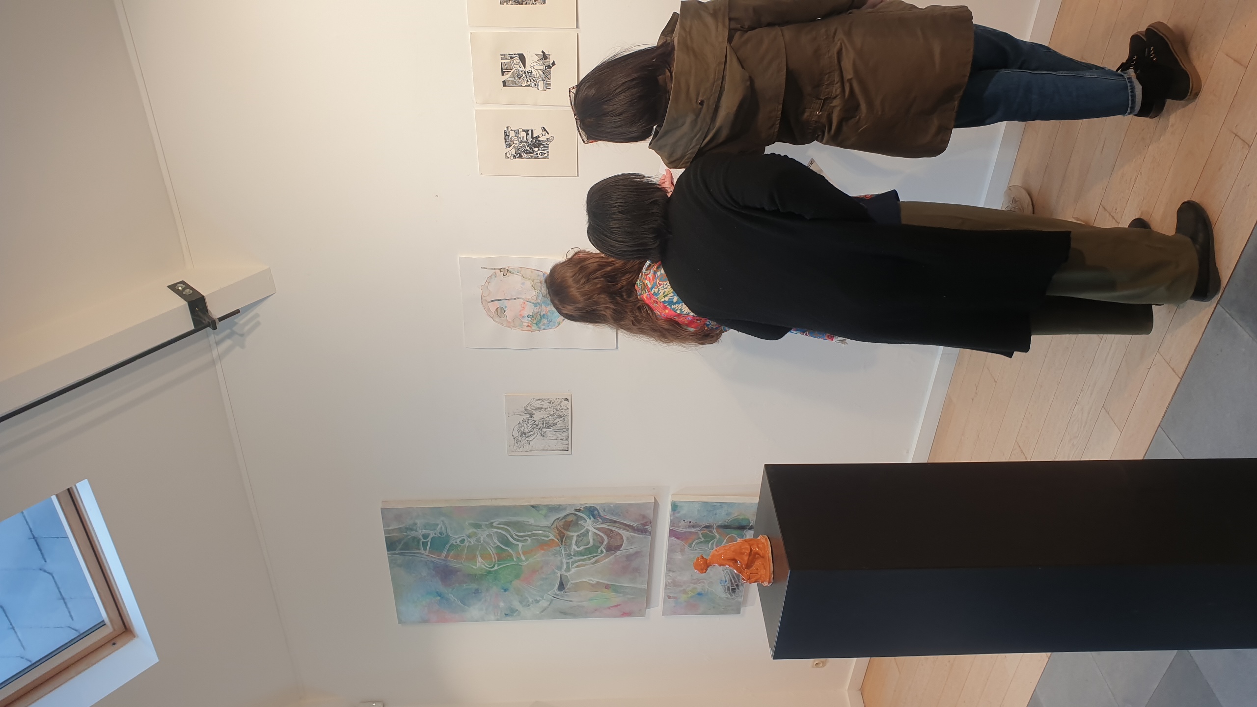 Trois personnes de dos visitent une exposition en galerie blanche, admirant des peintures colorées et des dessins encadrés au mur, avec une sculpture orange sur un socle noir au premier plan.