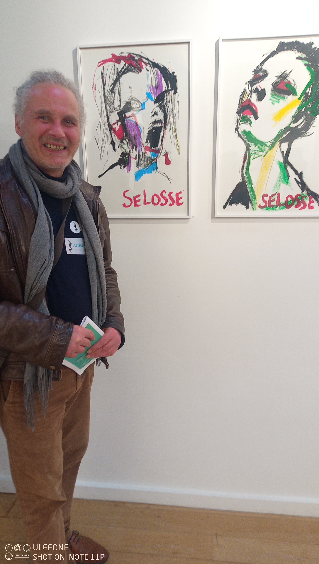 Un homme souriant pose devant deux portraits numériques abstraits signés Selosse, aux traits vigoureux et aux teintes vives de rose, bleu, vert et jaune, lors d'une exposition en galerie.