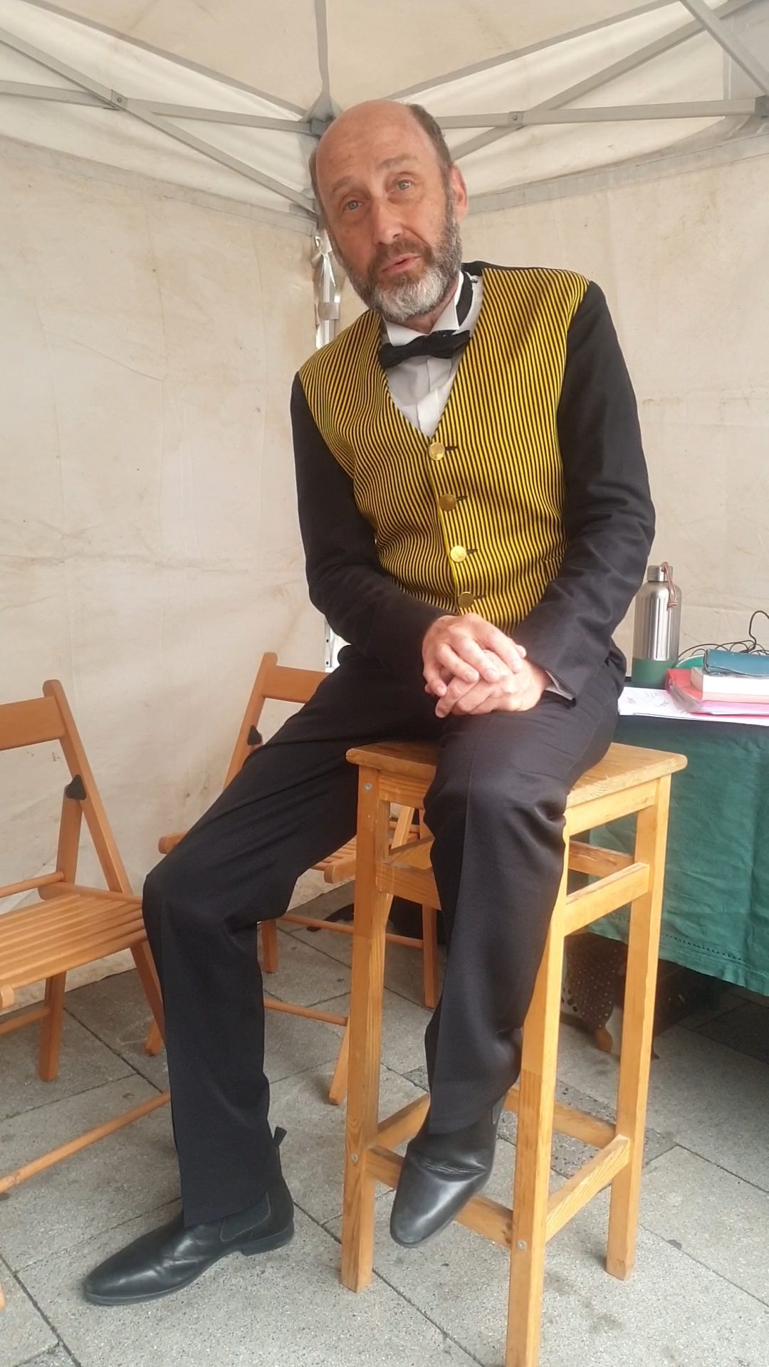 Bruno Georis en costume rayé jaune et nœud papillon, assis sous une tente lors d'une performance