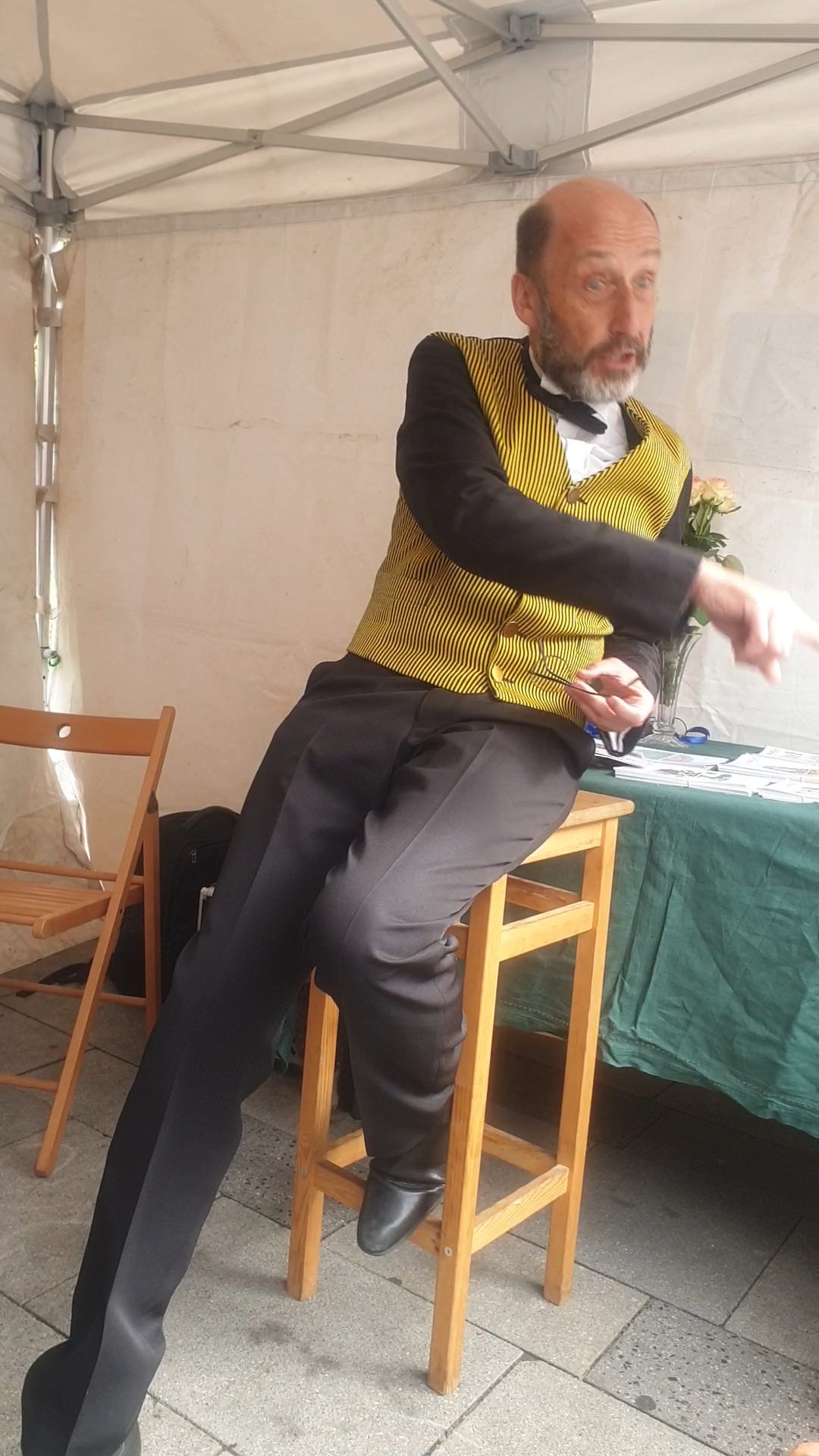 Bruno Georis - comédien et poète - vêtu d'un gilet jaune à rayures et d'une chemise blanche, assis sur un tabouret en bois, tient une fleur à la main et s'exprime sous un chapiteau blanc.